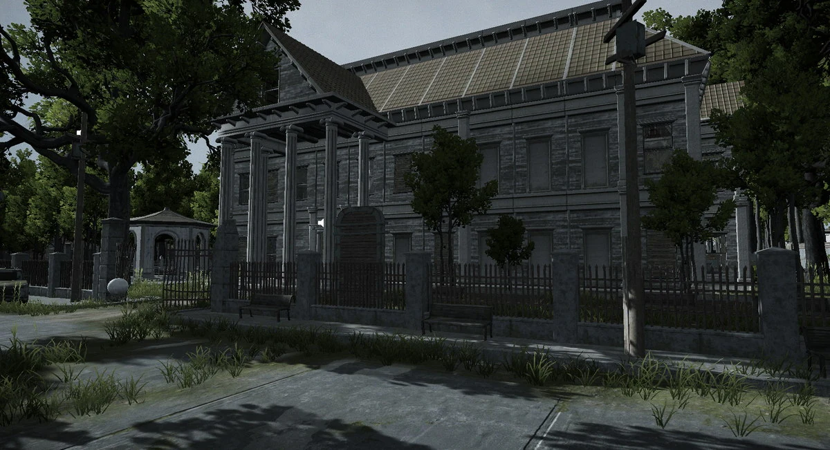 A large grey stone building with a colonnaded porch and boarded windows, standing behind iron railings with empty benches and overgrown grass in front